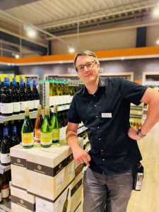 Ein Mann in schwarzem Kissel SBK Hemd und Brille steht lächelnd in der Weinabteilung eines Supermarkts. Er lehnt sich an einen Stapel Weinkartons mit aufgestellten Weinflaschen. Im Hintergrund sind Regale mit weiteren Weinflaschen zu sehen. Es handelt sich um ein Vorstellungsbild des neuen Marktleiters Herr Schwarzmüller im EDEKA Kissel SBK-Markt in Edenkoben.