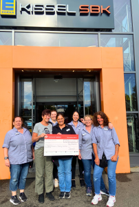 Eine Gruppe von sieben Frauen steht lächelnd vor dem Eingang des EDEKA Kissel SBK in Bad Bergzabern. Sie halten gemeinsam einen symbolischen Spendenscheck der Sparkasse Südpfalz über 1.000 Euro. Die Spende ging an die "Riding Santas" und wurde durch ein Fest in Bad Bergzabern gesammelt. Sechs der Frauen tragen EDEKA-Arbeitskleidung, eine Frau in der Mitte ist zivil gekleidet. Im Hintergrund ist der moderne Eingangsbereich des Supermarkts mit orangen Rahmen zu sehen.