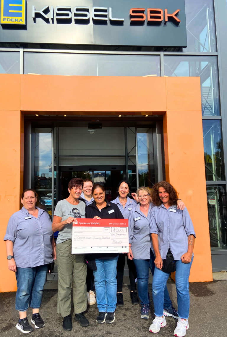 Eine Gruppe von sieben Frauen steht lächelnd vor dem Eingang des EDEKA Kissel SBK in Bad Bergzabern. Sie halten gemeinsam einen symbolischen Spendenscheck der Sparkasse Südpfalz über 1.000 Euro. Die Spende ging an die "Riding Santas" und wurde durch ein Fest in Bad Bergzabern gesammelt. Sechs der Frauen tragen EDEKA-Arbeitskleidung, eine Frau in der Mitte ist zivil gekleidet. Im Hintergrund ist der moderne Eingangsbereich des Supermarkts mit orangen Rahmen zu sehen.