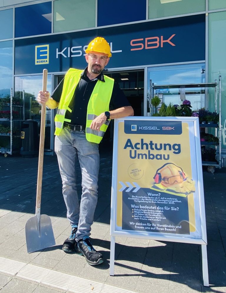 Unser Marktleiter aus Walldorf-West, steht vor dem Eingang des EDEKA Kissel SBK-Markts. Er trägt eine gelbe Warnweste und einen gelben Bauhelm und lehnt sich mit einem Arm an ein Baustellenschild mit der Aufschrift „Achtung Umbau“. In der anderen Hand hält er eine Schaufel. Im Hintergrund ist das Marktlogo sowie ein Regal mit Pflanzen zu sehen.