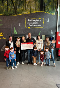 Zu sehen sind ist ein Mitarbeiter des Edeka Kissel SBK Pirmasens in Arbeitskleidung, sowie vier Frauen mit Kindern in Zivilkleidung. Der Mann und eine Frau stehen in der Mitte und halten einen großen Spendencheck der Sparkasse in den Händen. Das Foto ist entstanden bei der Spendenübergabe an einen Kindergarten im SBK-Markt in Pirmasens.