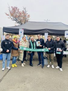 Zu sehen sind mehrere Mitglieder des Fußballvereins "TV 1896 Kirrweiler", die einen grün-weißen Schal vor sich halten auf dem steht "Reblandhölle". Zwei der sieben abgebildeten Personen zeigen ein Stickeralbum und ein Spielerbilder vor sich. Hinter der Gruppe ist ein überdachter Verkaufsstand aufgebaut, bei dem Glühwein, Waffeln, Kaffee und Fanartikel verkauft werden. Entstanden ist das Foto im Zuge der Stickerstar-Aktion des TV Kirrweiler in Zusammenarbeit mit EDEKA Kissel SBK.