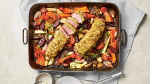 Zu sehen ist eine große, rechteckige Ofenform, die mit zwei rosa gegarten Lammfilets gefüllt ist, die mit einer goldbraunen Kräuterkruste bedeckt sind. Eines der Filets ist bereits in mehrere Scheiben geschnitten, sodass das saftige, zart rosafarbene Fleisch im Inneren gut sichtbar ist. Rund um das Fleisch liegen bunt gemischte, im Ofen geröstete Gemüsestücke, darunter rote Bete, Karotten, Pastinaken, kleine Tomaten, Schalotten und Rosenkohlhälften. Das Gemüse ist leicht karamellisiert und in kräftigen Rot-, Orange- und Gelbtönen gehalten. Die Form steht auf einem hellgrauen Tuch, daneben liegen ein Teller, eine kleine Schale mit heller Sauce und ein Messer.