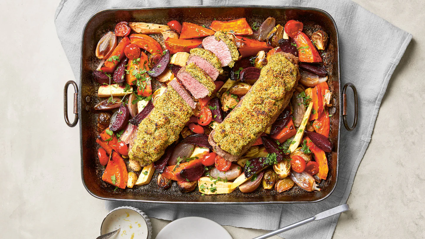 Zu sehen ist eine große, rechteckige Ofenform, die mit zwei rosa gegarten Lammfilets gefüllt ist, die mit einer goldbraunen Kräuterkruste bedeckt sind. Eines der Filets ist bereits in mehrere Scheiben geschnitten, sodass das saftige, zart rosafarbene Fleisch im Inneren gut sichtbar ist. Rund um das Fleisch liegen bunt gemischte, im Ofen geröstete Gemüsestücke, darunter rote Bete, Karotten, Pastinaken, kleine Tomaten, Schalotten und Rosenkohlhälften. Das Gemüse ist leicht karamellisiert und in kräftigen Rot-, Orange- und Gelbtönen gehalten. Die Form steht auf einem hellgrauen Tuch, daneben liegen ein Teller, eine kleine Schale mit heller Sauce und ein Messer.