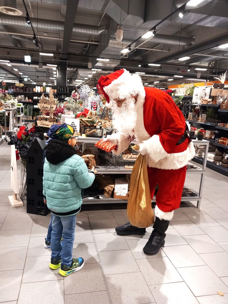Zu sehen ist ein Mann in rot-weißem Nikolauskostüm, der einem kleinen Jungen ein Geschenk überreicht. Der verkleidete Mann hält einen braunen Jutesack, befüllt mit weiteren kleinen Geschenken, in der Hand. Im Hintergrund zu erkennen ist die Dekorationsabteilung des Marktes in Bad Bergzabern.